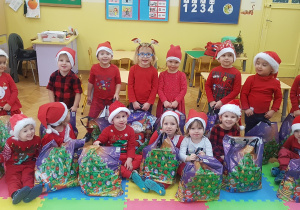 zdjęcie zbiorowe. Dzieci w czerwonych strojach i mikołajowych czapkach, trzymają w rączkach torby z prezentami od Mikołaja
